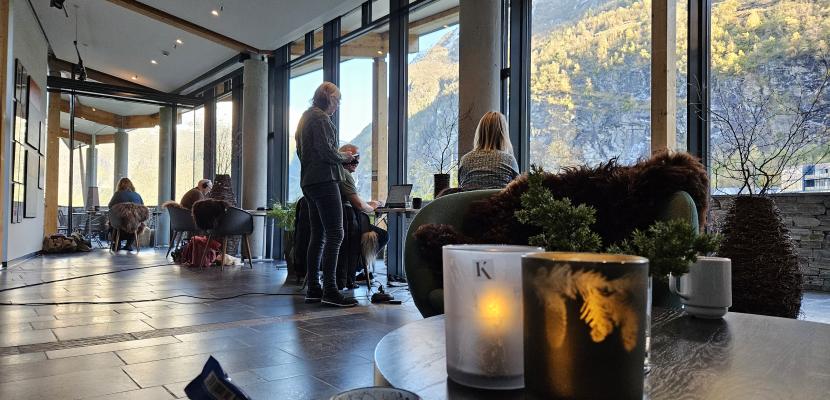 Cowork in Geiranger Photo of a cowork facilities with people sitting by a stretch of window. Outside there are mountains.