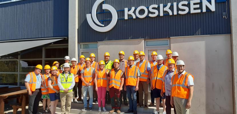 The Päijät-Häme Chairpersons’ Forum on a Company Visit Outdoor group photo from a factory visit, featuring around 30 people posing while wearing orange vests and yellow helmets.