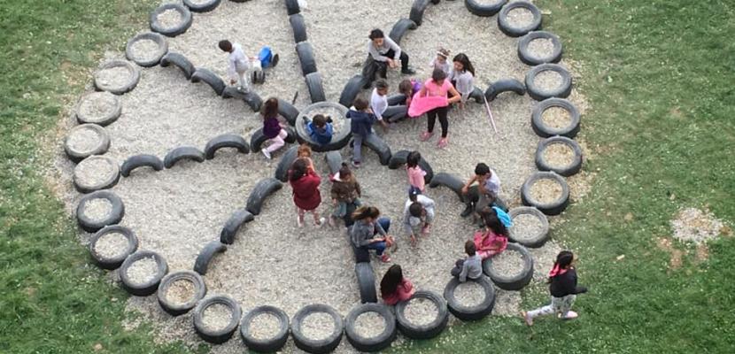 Children playing at the playground The image shows an outdoor play area where a group of children are gathered. The play area is designed in a circular pattern using old tires, which are arranged in a structured way—possibly resembling a flower or a peace sign. The inside of the structure is filled with gravel or small stones. The children are actively engaging with the space, some sitting on the tires while others stand or walk around. The setting appears to be a grassy field, and the atmosphere is lively and interactive.