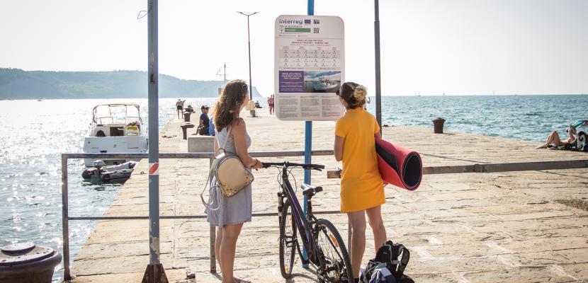 The image shows cyclists waiting for a boat, chacking the timetable. The boat provides transport for passengers and bicycles, complementing the offer for cyclists.