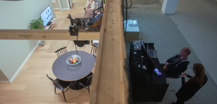 Inside and Outside: Observing the Test Apartment in Action The image shows the test apartment viewed from above, with two people sitting inside the apartment and having a conversation. Outside the apartment, two people are observing the activity inside on monitors.