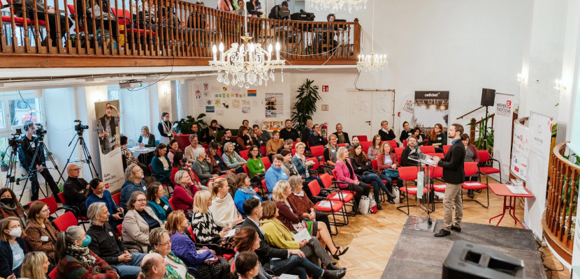WienZimmer Gartenstadt – Grätzlzentrum (Community Center) des Fonds Soziales Wien Conference at Grätzlzentrum (Community Center) of the Fonds Soziales Wien. A speaker stands on a small stage, addressing an audience seated on red chairs. A balcony above hosts more participants. Cameras on tripods record the event.