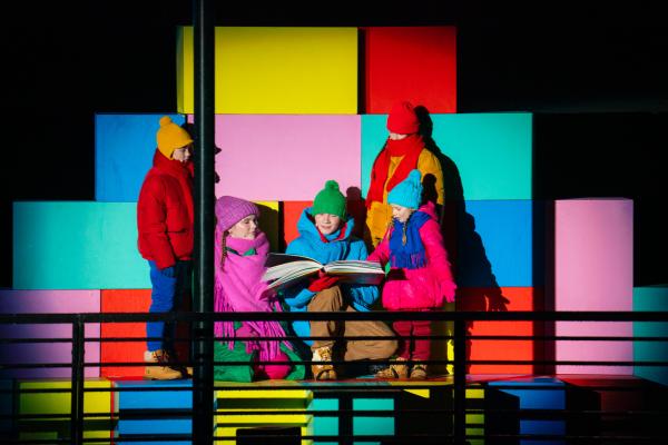 Five children and young people in colorful clothing reading large book on stage