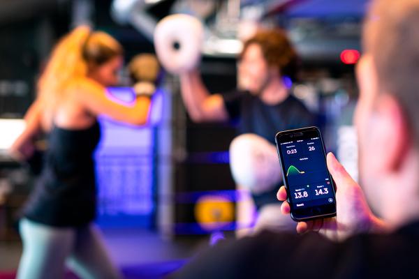 In front a man holding a phone showing data. In the backround two women are boxing.