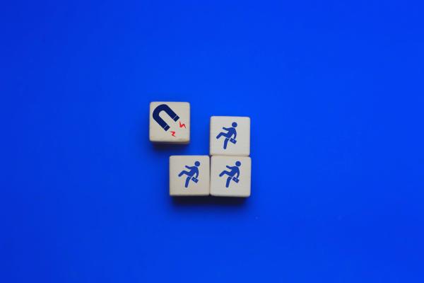 Four wooden blocks are arranged on a bright blue background. One block features a magnet icon with red lines indicating attraction, while the other three blocks each display a businessperson figure walking toward the magnet.