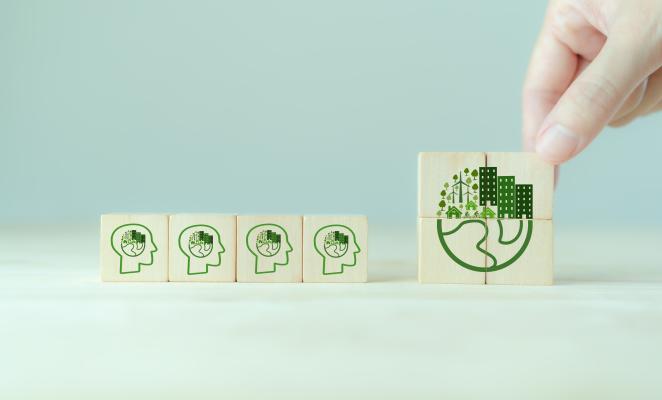 The image shows a close-up of a hand holding a rectangular wooden block. The block has a flat, colorful illustration glued to the top surface. The illustration depicts a globe formed from a cityscape with skyscrapers and other buildings.