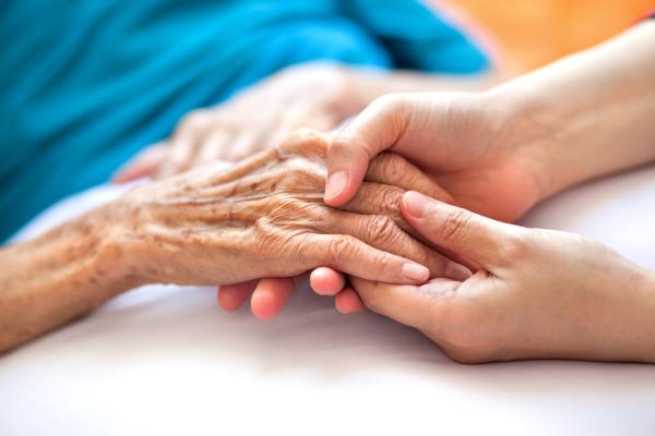 The image depicts a close-up of two hands: one is older, showing visible wrinkles and age spots, and the other is younger, gently holding the older hand. In the blurred background, a person is lying down, covered with a blue blanket or garment. This scene conveys a sense of care and connection between generations.