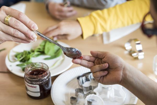 A close-up of two hands during a shared cooking activity—one adult holds a spoon with homemade jam, gently placing a small amount onto a child's fingertip.