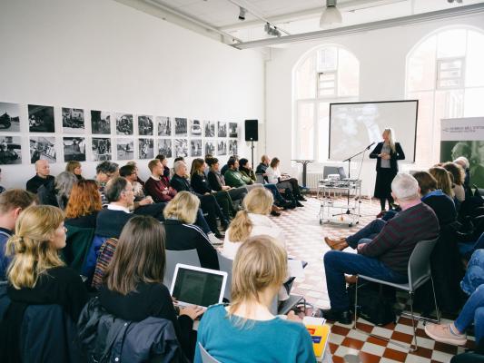 A group of people sitting in a room in an u-shape, a woman holds a speech
