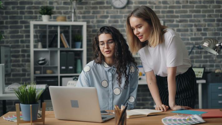 Two women collaborate at a desk in front of a computer