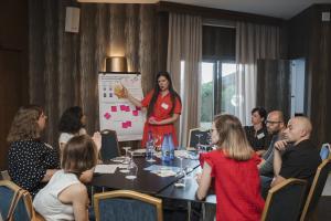 Woman dressed in red standing in front of flipchart presenting to group of people sitting around a table