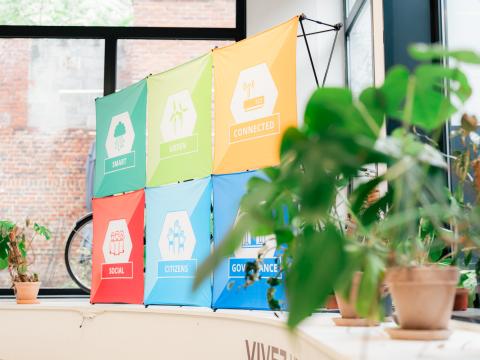 Projects banner with green plant Six colorful Interreg Europe project logos are positioned in a printed banner, sitting in front of a window in an industrial corporate space. A plant sits in the foreground of the image, closest to the camera.