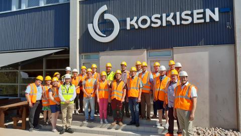 The Päijät-Häme Chairpersons’ Forum on a Company Visit Outdoor group photo from a factory visit, featuring around 30 people posing while wearing orange vests and yellow helmets.