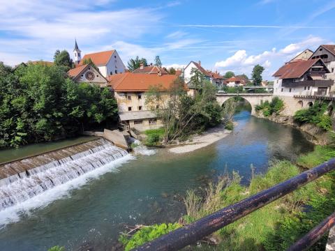The charming town of Škofja Loka, Slovenia, provided a serene and inspiring setting for the FBI4EU project kickoff meeting, fostering collaboration amidst its historic architecture and natural beauty. A scenic view of a waterfall flowing into a river in Škofja Loka, Slovenia, with historic buildings and a stone bridge in the background. The red-tiled roofs and lush greenery complement the serene blue sky, creating a picturesque landscape.