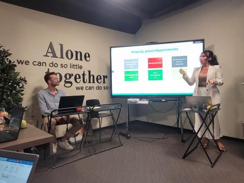 During the FBI4EU project kickoff meeting, partners engaged in a detailed discussion about priority areas and departments, emphasizing the importance of collaboration for successful regional development. A woman stands presenting in front of a large screen displaying priority areas and departments, while a man sits nearby with a laptop. The room features a motivational quote on the wall that reads, "Alone we can do so little; together we can do so much.