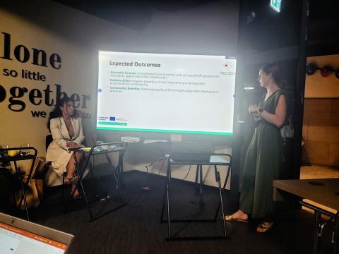 During the FBI4EU project kickoff meeting, partners presented their policy instruments, discussing expected outcomes such as economic growth, sustainability, and community benefits, crucial for driving regional development. Two women are presenting in a conference room. One is standing and speaking in front of a screen displaying "Expected Outcomes," while the other is seated, listening attentively. The wall features a motivational quote that reads, "Alone we can do so little; together we can do so much."