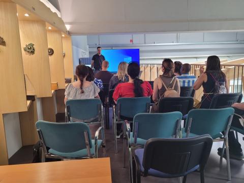 FBI4EU project participants attending an informative session at MSora, learning about the company's innovative approaches and sustainable practices in the wood industry. A group of people seated in a room with wooden displays, listening to a presentation by a speaker standing in front of a large screen.