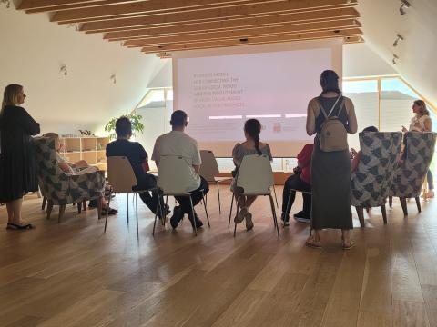 Participants of the FBI4EU project engaged in a session discussing business models for utilizing local wood and developing high-value-added wood products, highlighting innovative strategies for regional development. A group of people seated in a well-lit room with wooden floors and ceiling beams, watching a presentation on a large screen about the business model for connecting the use of local wood and developing high-value-added wood products.