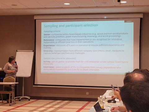 LRCAF Presentation on Good Practices Surveys and Interviews A presenter standing near a screen displaying a slide titled "Sampling and participant selection." The slide outlines criteria such as sector, relevance, experience, and diversity for selecting companies in forest-based industries, along with survey and interview goals.