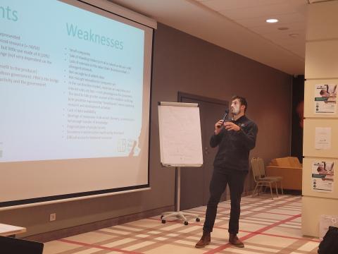 Presentation of SWOT Analysis by Filière Bois Wallonie A presenter standing near a screen displaying a slide with the title "Weaknesses," part of a SWOT analysis. The slide lists challenges faced by forest-based industries, such as lack of cohesion, low added value, and data availability issues. A flipchart is visible nearby. Title: Presentation of SWOT Analysis by Filière Bois Wallonie