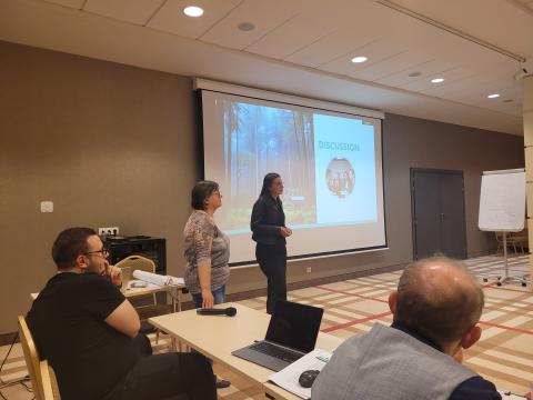 Methodology for Preparing a Joint Report on the Forest-Based Industry's Current Situation Two presenters stand near a screen displaying a slide with the title "Discussion" and an image of a forest and a group of people at a table. The presentation appears to engage the audience, some of whom are seated and taking notes.