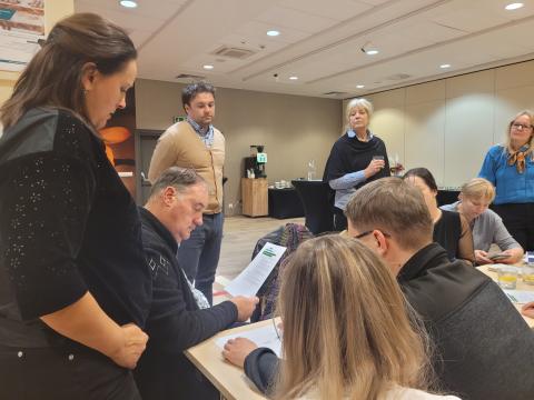 Workshop Session During the FBI4EU Project Meeting A group of participants actively engaged in a workshop discussion, with some seated and reviewing documents while others stand nearby observing or contributing to the conversation. The setting is a conference room.