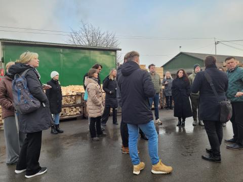 Study Visit to Drew BIK A group of individuals in warm clothing stand outdoors during a site visit, with piles of wood and a green warehouse visible in the background. The participants appear engaged in conversation or listening to a guide.