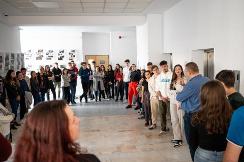 A group of young people stands in a hall of an educational institution, while a man in a blue shirt speaks to them.