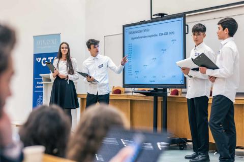 A group of young students in formal attire presents financial data on a large screen in a classroom. One student points at the chart, while others hold folders. The audience listens attentively.