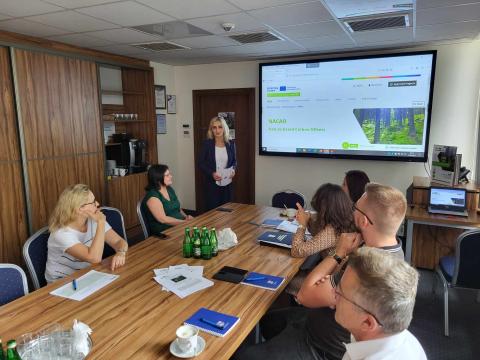 Third stakeholder meeting in Poland August 2024 Group of people seated around a conference table, listening to a woman presenting. A large screen in the background displays the presentation.