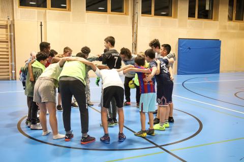 A group of boys, wearing sports gear and indoor football shoes, are standing in a team huddle, inside a sports hall. The goal keeper is standing in the middle of the circle. There is one adult. 