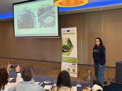 A woman stands in front of a projected presentation slide at a conference, speaking to an audience. The slide displays two labeled images: one of manure and one of cheese whey, under the topic of sustainable resource use. Next to her is a vertical banner for the "3F GREEN MODEL" project, co-funded by the European Union under Interreg Europe, promoting circular economy policies. Audience members are seated at tables, with one person in the foreground photographing the speaker with a phone.