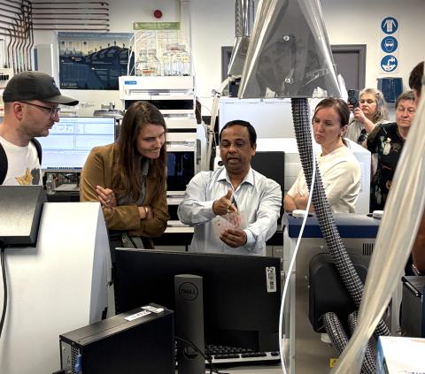 A group of people gather in a laboratory around a researcher who is explaining a sample, which he holds in his hands. The lab is filled with advanced analytical equipment, computer monitors, and ventilation hoods. Some participants listen attentively while others observe quietly in the background, with one person recording the scene on a phone. The environment is modern and technical, emphasizing a hands-on demonstration or tour focused on scientific instrumentation or analysis.