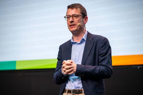 Nicolas Singer Nicolas Singer stands on stage during a professional event wearing a suit and a microphone attachment. He is speaking to a crowd.