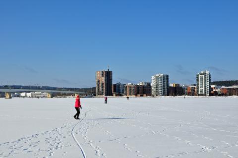 Forzen lake, two people walking on ice and a city background on a shore.