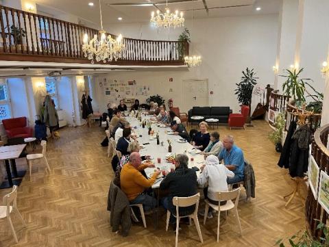 A Long table placed diagonally within a large room. People are seated alongside, with plates of food in front of them. 