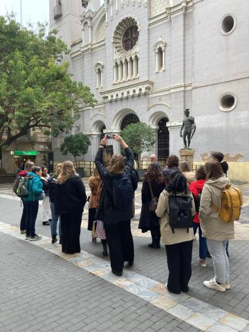 JUSTGREEN Murcia city walk The group is outside on a classic Spanish square. In the left corner part of an ancient tree is visible while the group is standing in front of a church like building with a mid-size statue in front. One person is taking a picture while the rest is listening to an explanation.