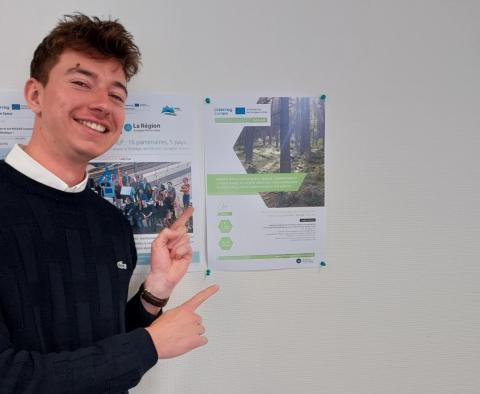 Yann Mathias, dark-haired studen pointing to the NACAO poster and smiling