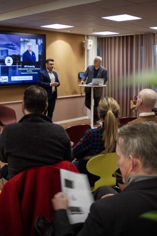 Stakeholder meeting Two speakers stand at the front of a conference room, addressing an audience seated in colorful chairs. A presentation slide is displayed on a large screen.
