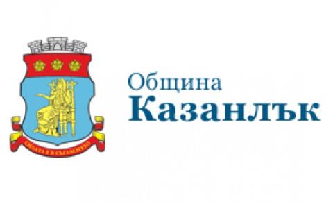 Municipality of Kazanlak A full-color heraldic shield depicting the coat of arms of Kazanlak, Bulgaria. The upper field is red and bears three stylized golden rosebuds in a horizontal row. The lower, larger field is blue and shows a golden female figure seated on a throne, holding a distaff and spindle, symbolizing the region’s historic rose oil and textile traditions. Above the shield sits a silver mural crown with three visible towers. Beneath the shield, a red ribbon bears the town’s motto in gold Cyrillic lettering: “Силата е в