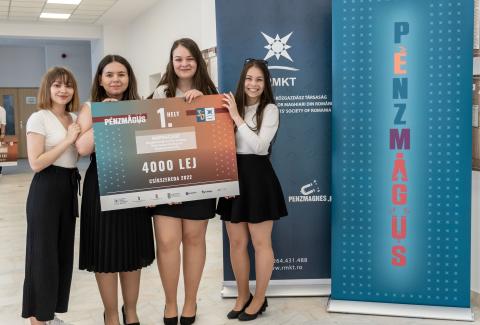 Four young students stand together indoors, holding a large check for 4000 RON, awarded for winning the Money Magician competition. They are smiling, dressed in semi-formal attire, with roll-up banners behind them.