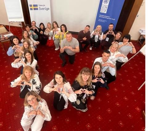People sit grouped in a heart shape, making heart symbols with their hands
