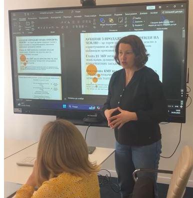 A woman stands in front of a video screen, talking to the attendees