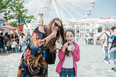 A child and an adult eating ice cream in front of a Ferris Wheel. 