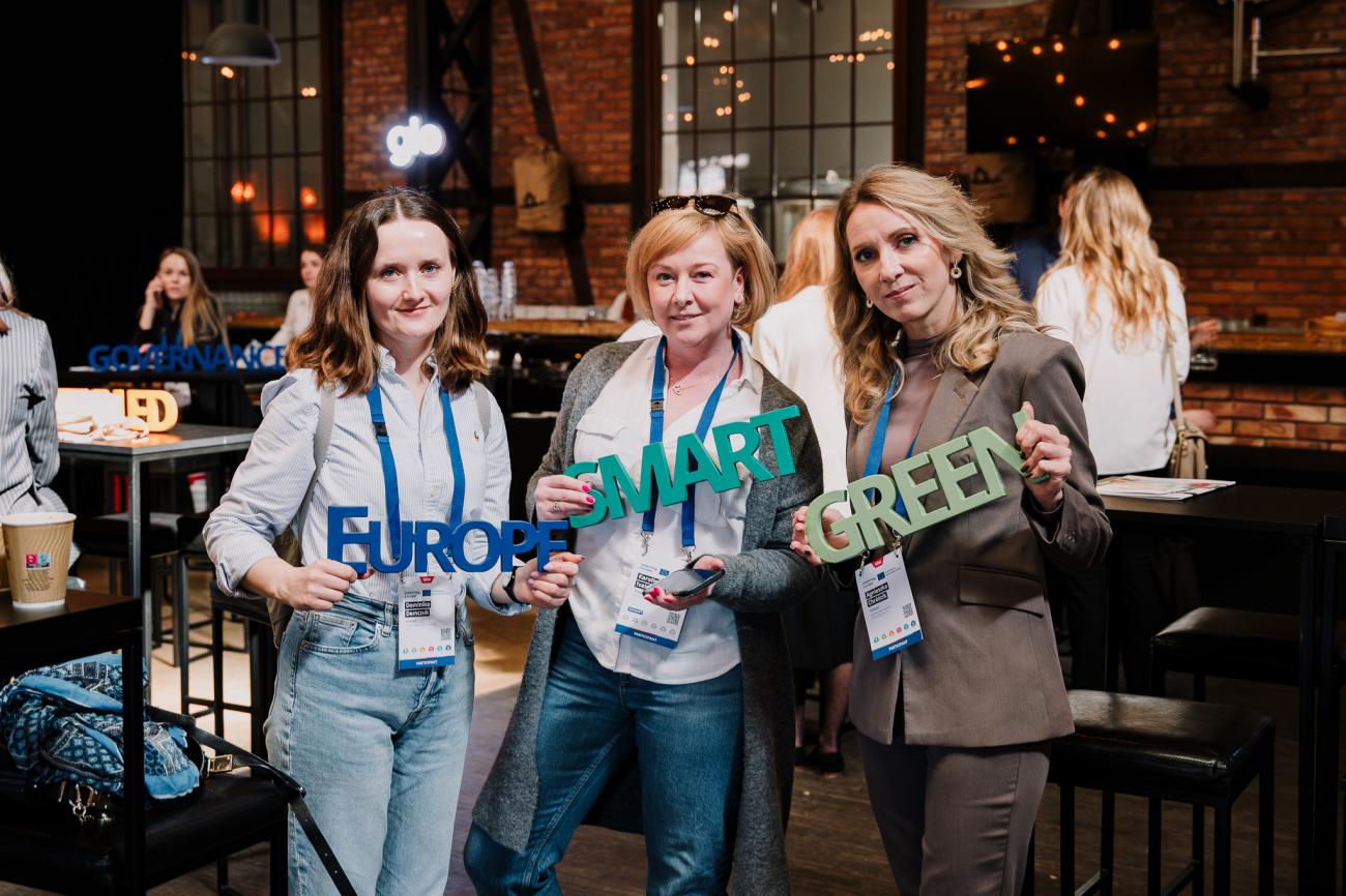Three women pose with wooden words for the camera at Europe, let's cooperate 2025 in Krakow
