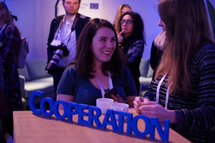Two people talk and laugh during an event standing next to a table with the word cooperate on it