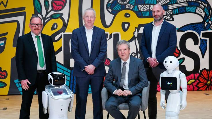 The image shows four men in suits, two standing and two seated, along with two humanoid robots.