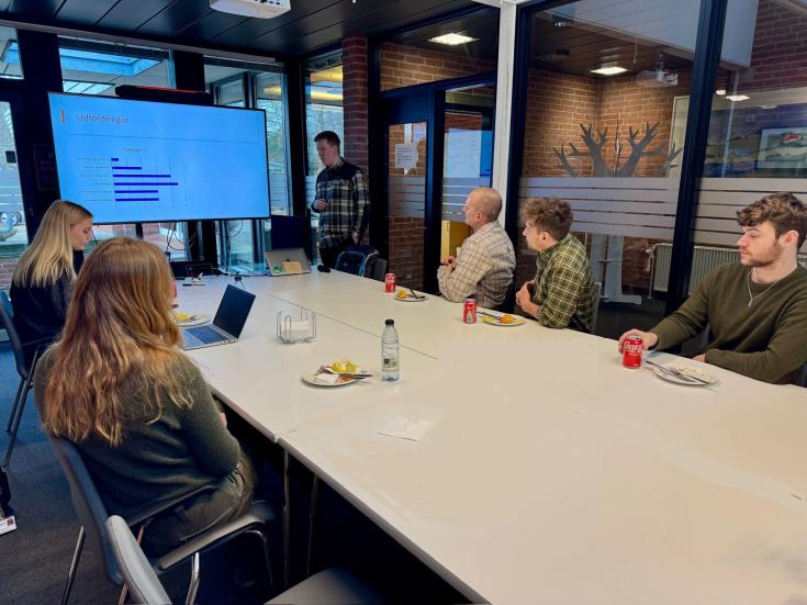 Stakeholder Meeting Presentation on Forestry Challenges A group of people is attending a presentation in a modern meeting room with large windows. A presenter stands near a screen displaying a bar chart labeled "Udfordringer" (Challenges). Participants sit around a table, listening, eating, and taking notes.