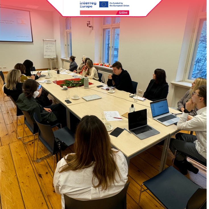 4th Regional Stakeholder Group Meeting - Latvia Meeting participants seated around tables arranged together. All looking towards the screen on which the presentation is projected. In the corner of the room to the right of the screen, a whiteboard is visible.