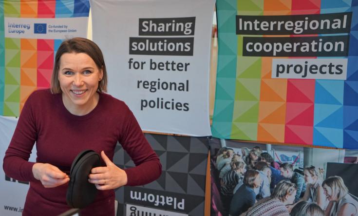 person smiling and standing in front of a colourful Interreg Europe stand and holding headphones